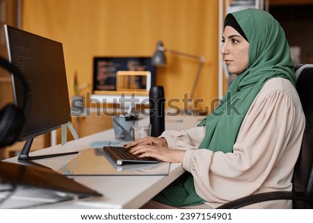 Side view portrait of successful Muslim woman as female programmer writing code in office and wearing modest clothing