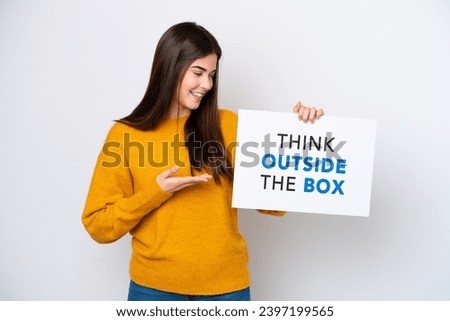 Young Brazilian woman isolated on white background holding a placard with text Think Outside The Box and  pointing it
