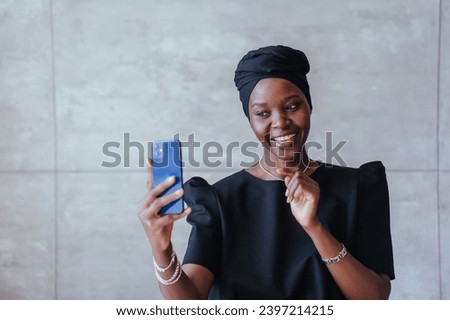 Joyful African girl in black turban talks by phone makes video call against marble wall with copy space.Brazilian young woman streaming via internet. Blogger.
