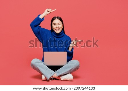 Full body young IT woman of Asian ethnicity wear blue sweater casual clothes sitting work hold use laptop pc computer point aside on area isolated on plain pastel pink background. Lifestyle concept