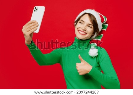 Smiling young woman wear green turtleneck Santa hat posing do selfie shot mobile cell phone show thumb up isolated on plain red background. Happy New Year 2024 celebration Christmas holiday concept