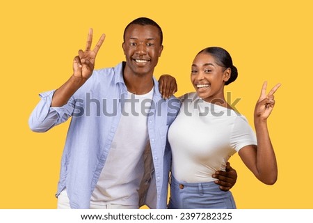 Radiant young black couple smiling broadly, making peace signs with their hands against monochrome yellow background, smiling at camera