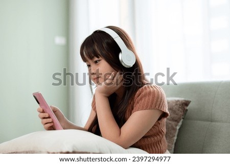 Young cute Asian girl in casual clothes wearing headphones and watching kid cartoons online on her smartphone on a sofa in the living room. Kid and technology concepts
