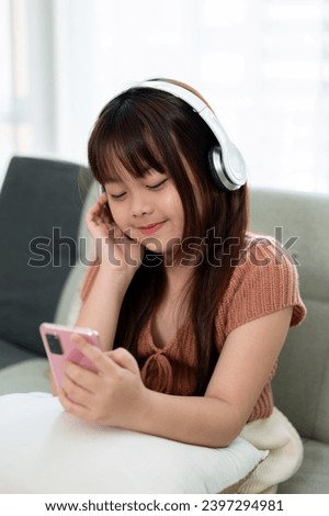 Young adorable Asian girl in casual clothes wearing headphones and using her smartphone, enjoying listening to music on a sofa in the living room. Kid and technology concepts
