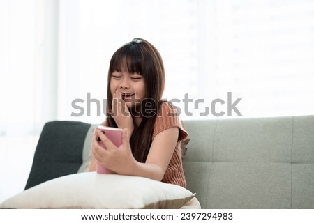 Young cute Asian girl in casual clothes using her smartphone on a sofa in a living room, relaxing and watching kid cartoons on a smartphone at home. Kid and technology concepts