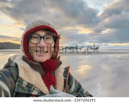 Mature woman wearing warm clothing takes a selfie for social media while walking on the beach on Rhossili Beach in Wales at sunset.