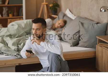 Man with smartphone in bedroom, woman sleeping in the background Royalty-Free Stock Photo #2397310061