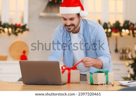Celebrating Christmas online with exchanged by mail presents. Smiling man in Santa hat opening gift box during video call on laptop at home