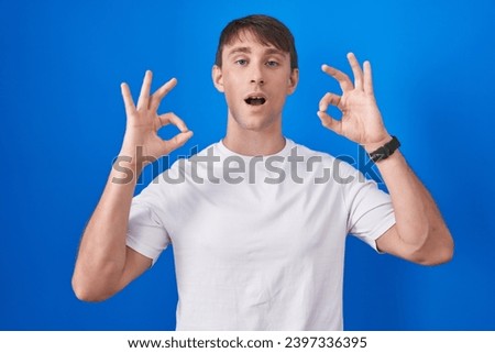 Caucasian blond man standing over blue background looking surprised and shocked doing ok approval symbol with fingers. crazy expression 