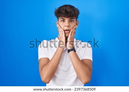 Hispanic teenager standing over blue background afraid and shocked, surprise and amazed expression with hands on face  Royalty-Free Stock Photo #2397368509