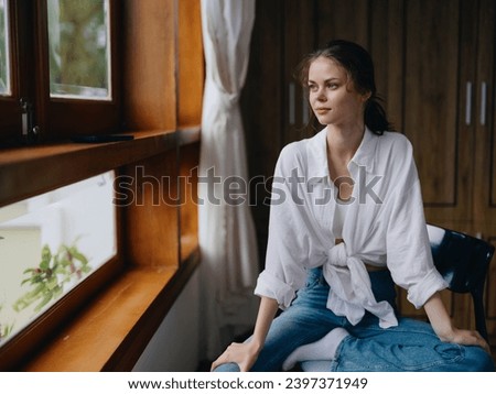 Woman sitting at home by a wooden window with a smile on a chair in homemade comfortable clothes and looking at the landscape, spring mood, women's day, rest on the weekend. Royalty-Free Stock Photo #2397371949