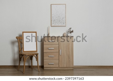 Wooden chair, chest of drawers, decorative elements and frames in room with light wall, space for text. Interior design