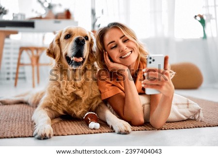 Saving memories with pet. Smiling woman with blond hair snuggling to furry friend and taking selfie on modern cell phone. Obedient golden retriever lying on floor near delighted female owner. Royalty-Free Stock Photo #2397404043