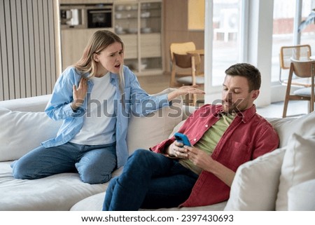 Indifferent ignoring man sits on sofa with mobile phone, playing games, scrolling social networks, reading news, keeping silence while offended irritated woman raises voice and shouts reproachfully Royalty-Free Stock Photo #2397430693