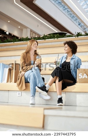 Two biracial friends sitting in rest area at mall talking and drinking lemonade Royalty-Free Stock Photo #2397443795