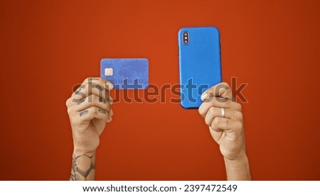 Hispanic man's tattooed hands holding phone over isolated red wall, texting payment message, male's online shopping spree magnifying his financial debt.