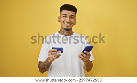 Happy, confident young latin man, casually standing, engages in online shopping using his smartphone and credit card over the vibrant yellow isolated background