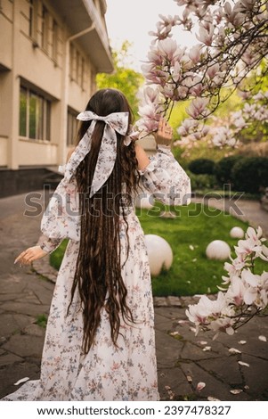 Brunette girl with long hair with big bow in long dress with train standing in garden of blooming magnolias in spring
