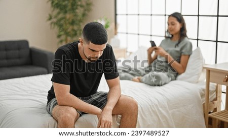 Beautiful couple disagreeing in their bedroom, sitting on the bed, expressing conflict while using a smartphone, a problem causing a serious expression