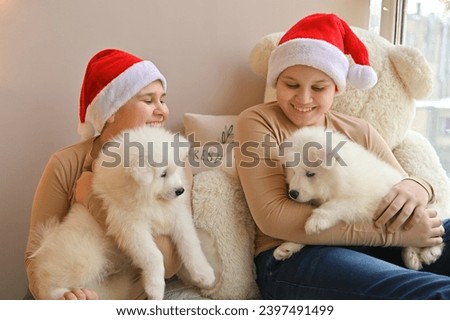 Christmas at home. Children and Samoyed puppies under the Christmas tree. Small children in a Santa hat hug a pet and open Christmas presents. Children are playing with an animal. 