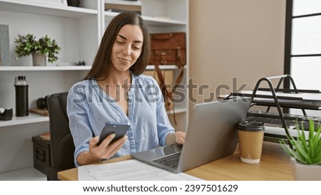 Attractive young hispanic woman immersed in success, confidently texting on her smartphone, typing work messages on her laptop, bossing it beautifully in the office