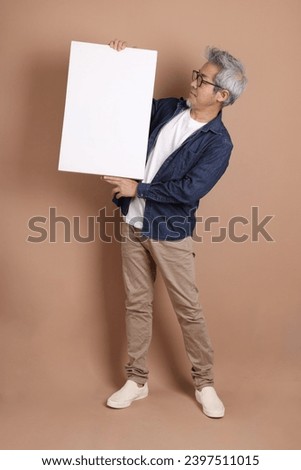 Asian energetic senior man with glasses holding a blank canvas in his hands  isolated on brown background with casual clothes. mature man, Asian man. Elderly,  studio shot. Multiple emotions. 