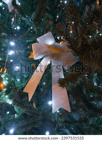 A beautiful bow made of silver ribbon is attached to a branch of the Christmas tree. Electric garland lights are burning around



