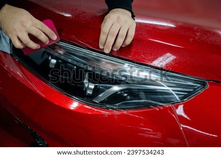 The master applies vinyl film to the headlight of a red car. Closeup view on worker detailer hand smoothing with a scraper protective film.  Royalty-Free Stock Photo #2397534243
