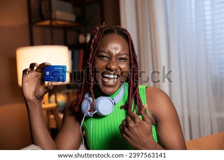 Portait of delighted cheerful afro american woman with colorful bright hair smiling looking at camera raising fist showing credit card at camera wearing modern new purple headphones sitting at home.