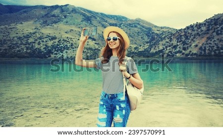 Summer vacation, young woman tourist taking selfie with mobile phone wearing straw hat with backpack on lake and mountains background. Greece, island Crete