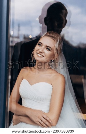 Beautiful bride near the window, reflection of the groom's silhouette. Wedding portrait of a cute blonde girl. Photography and concept.