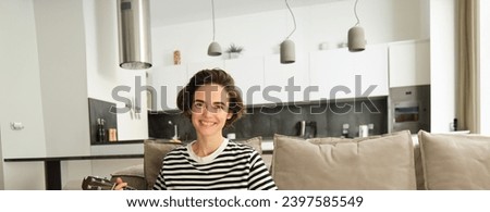 Vertical shot of young musician, woman learns how to play ukulele, sits on sofa with crossed legs and smiling at camera.