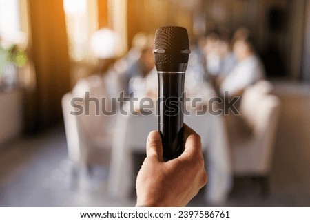 Unrecognisable male hand holds black microphone, master of ceremony or showman speech to audience in event. On background sit people guests by the table. Party and festive events concept