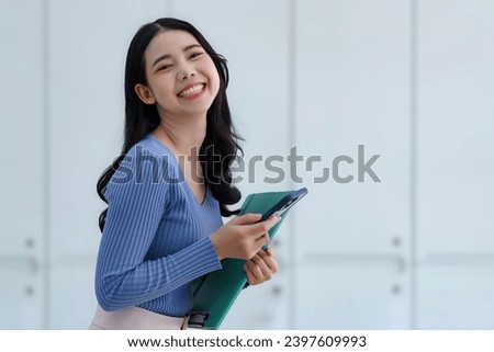 Young beautiful Asian business woman holding a folder and a smart phone while standing at the workplace.