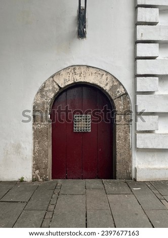 Medieval styled red door with round arch and small square metal grid window isolated on white wall. Outdoor european old age styled exterior architecture photography isolated on vertical ratio image.