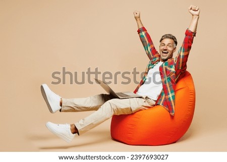 Full body adult IT man wear red shirt white t-shirt casual clothes sit in bag chair hold use work on laptop pc computer do winner gesture isolated on plain pastel beige background. Lifestyle concept