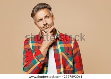 Adult man wear red shirt white t-shirt casual clothes put hand prop up on chin, lost in thought and conjectures isolated on plain pastel light beige color background studio portrait. Lifestyle concept