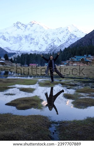 Asian female tourist having a picture taken with a view of tall mountain ranges with a snow capped mountains and its reflection in the pond on Fairy Meadows - a popular destination in Pakistan.