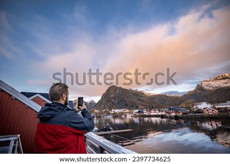 man taking a picture with phone of a rainbow appearing behind the top of a mountain over nordic Sørvågen village on lofoten islands in norway
