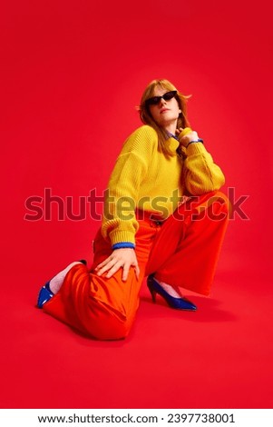 Cool attractive girl, student sitting posing dressed retro casual outfit in eyewear against vivid red background. Concept of bright colors in fashion ,human emotions, lifestyle, style trends.