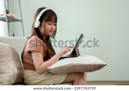 Young adorable and happy Asian girl in casual clothes wearing headphones and enjoying playing games or watching kid cartoons online on her digital tablet on a sofa in the living room.