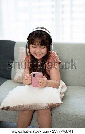 Young cute Asian girl in casual clothes wearing headphones and watching kid cartoons online on her smartphone on a sofa in the living room. Kid and technology concepts
