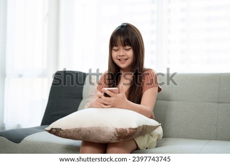 Young cute Asian girl in casual clothes using her smartphone on a sofa in a living room, relaxing and watching kid cartoons on a smartphone at home. Kid and technology concepts