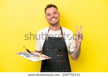 Young artist caucasian man holding a palette isolated on yellow background smiling and showing victory sign