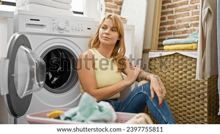 Young blonde woman sitting on floor leaning on washing machine tired at laundry room