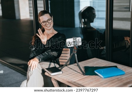 Female blogger gestures hello to subscribers at social media stream sitting at cafe outside. Pretty girl in casual makes video call talks to parents, relocation, new life.