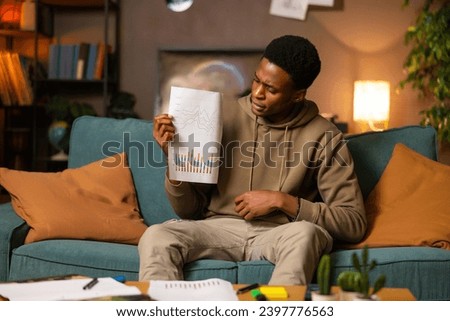 Sad exhausted african american dark skinned student tired of studying sitting in the living room showing at camera observing papers with graphics difficult study time at University exams time.
