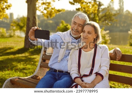 Happy old senior couple enjoying good sunny summer day relaxing on bench in beautiful park garden with green trees in background, using modern smart cell phone to take selfie with romantic life moment
