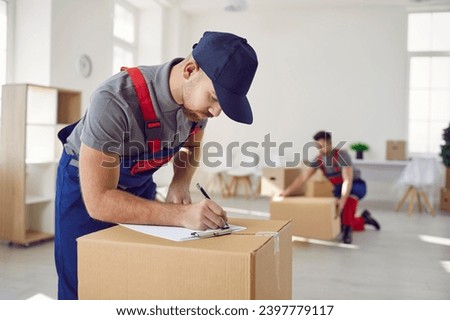 Worker from professional man and van delivery company writes something on paper while removing stuff on house moving day. Loader in uniform workwear and baseball cap fills out invoice on cardboard box Royalty-Free Stock Photo #2397779117