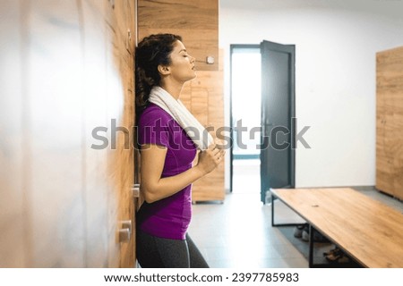 Tired athletic woman standing in locker room and taking a break from exercising. Woman holding gym towel while leaning against the locker in gym.